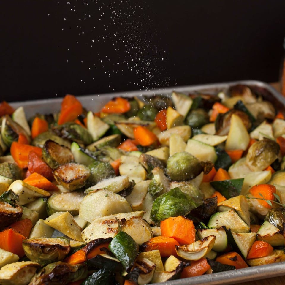 Salt sprinkles over a sheet pan of roasted vegetables, including carrots, zucchini, and Brussels sprouts.